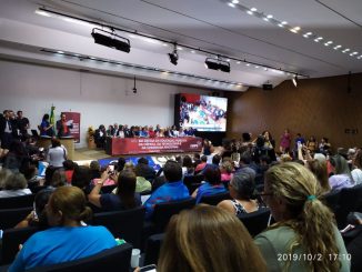 Várias pessoas assistem a um discurso em uma sala no congresso nacional. Na mesa está uma faixa vermelha escrito: ato em defesa da educação pública, da ciência, da tecnologia, e da soberania nacional.