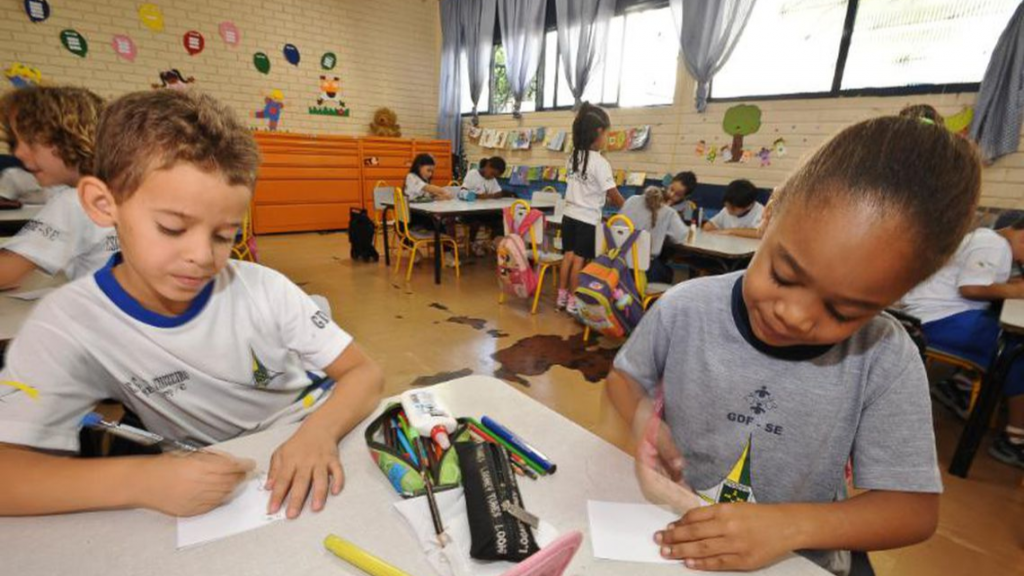 Duas crianças brincam com lápis de cor em uma mesa. Ao fundo, é possível ver uma sala de aula do Ensino Infantil com outras crianças.