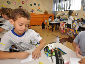 Duas crianças brincam com lápis de cor em uma mesa. Ao fundo, é possível ver uma sala de aula do Ensino Infantil com outras crianças.