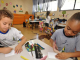 Um menino e uma menina brincam com lápis de cor. Sob a mesa, estão estojos e outros materiais escolares. No plano de fundo, é possível ver crianças e o interior de uma sala de aula de ensino infantil.