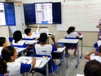 Crianças sentadas em uma sala de aula do ensino básico. Uma professora está à frente, próxima à lousa