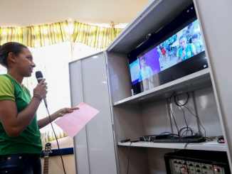 Na foto uma estudante em pé falando ao microfone com o professor que aparece na tela do monitor. Ensino à distância.