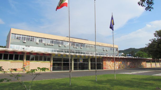 Foto da fachada frontal do prédio da reitoria da Universidade Federal de Santa Catarina À frente, há três bandeiras, da esquerda para a direita: bandeira de Santa Catarina, bandeira do Brasil e bandeira da UFSC
